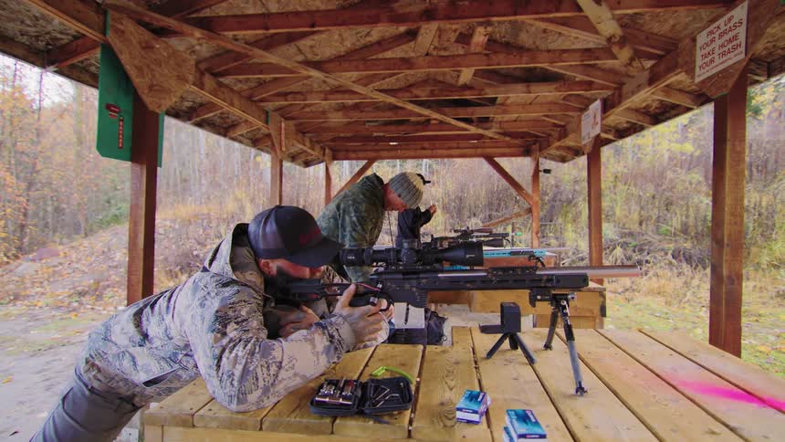 Men in camouflage practice their aim with high-powered rifles at a covered outdoor shooting range
