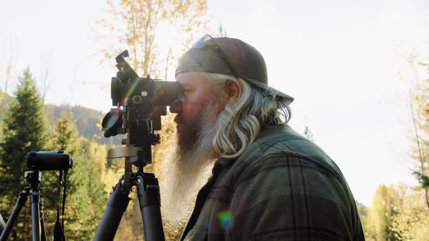 A bearded outdoorsman uses a spotting scope on a tripod while observing nature in a sun-drenched autumn forest