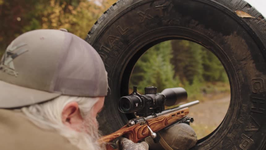 Close-up of a man with a white beard adjusting the scope on a rifle stabilized by a sandbag and framed by a large tire