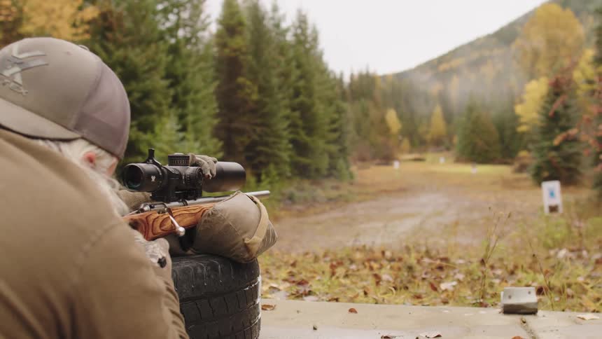 Slow motion over-the-shoulder shot of a man firing a bolt-action rifle at a scenic outdoor shooting range in autumn