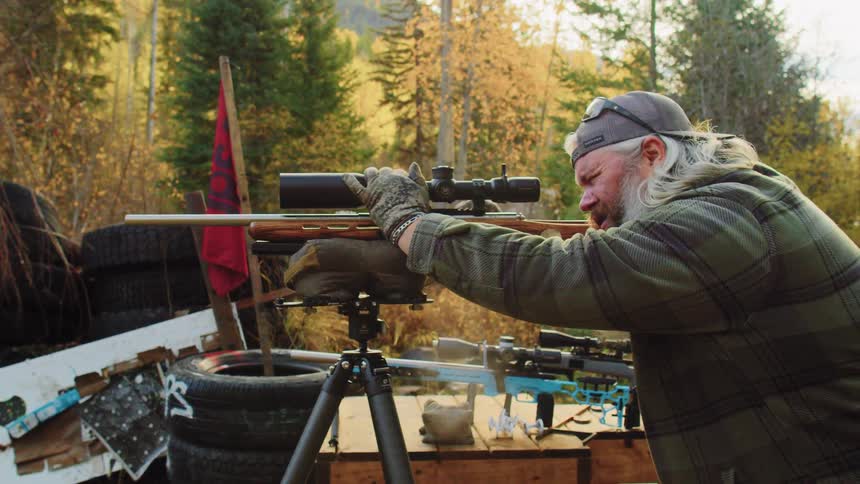 An experienced shooter in a plaid shirt and gloves takes careful aim with his scoped rifle on a tripod at a rustic outdoor range
