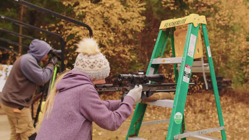 A woman in a winter hat fires a high-powered rifle off a ladder rest while a man spots for her during target practice