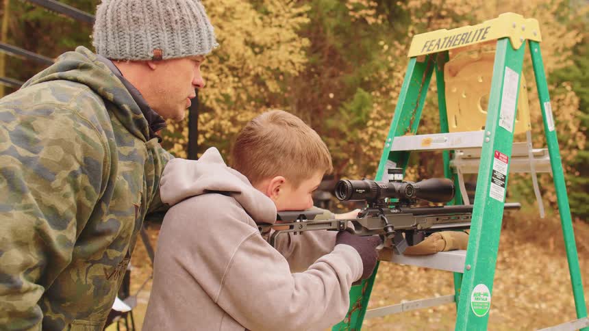 A father carefully guides his young son on how to safely aim and shoot a rifle at an outdoor target range