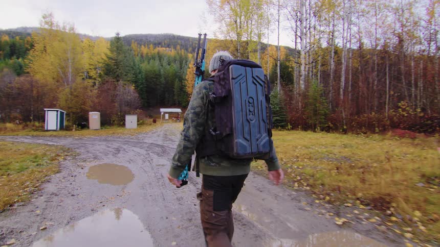 A man carrying a compound bow and a large backpack walks in slow motion down a muddy trail during autumn