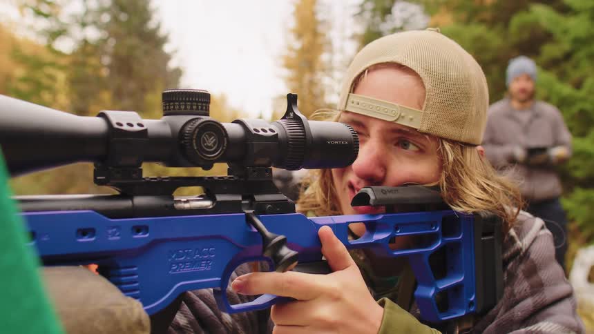 A focused shooter fires a blue bolt-action rifle, with the spent casing ejecting in slow motion
