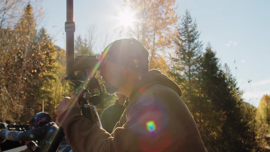 A man looks through a spotting scope at an outdoor range, with a shooter in the background on a sunny day