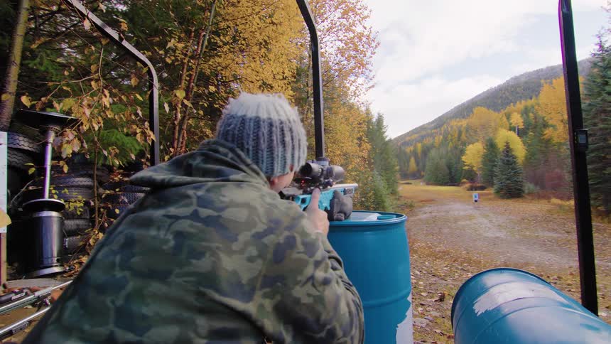 A man in camouflage shoots a scoped rifle at an outdoor range amidst autumn foliage