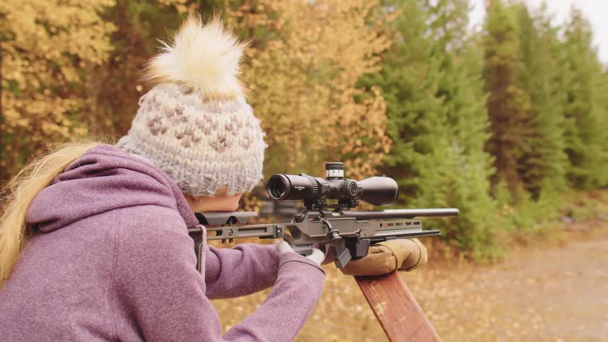 A young blonde woman in a winter hat adjusts her sniper rifle scope and takes aim at an outdoor shooting range in the autumn woods