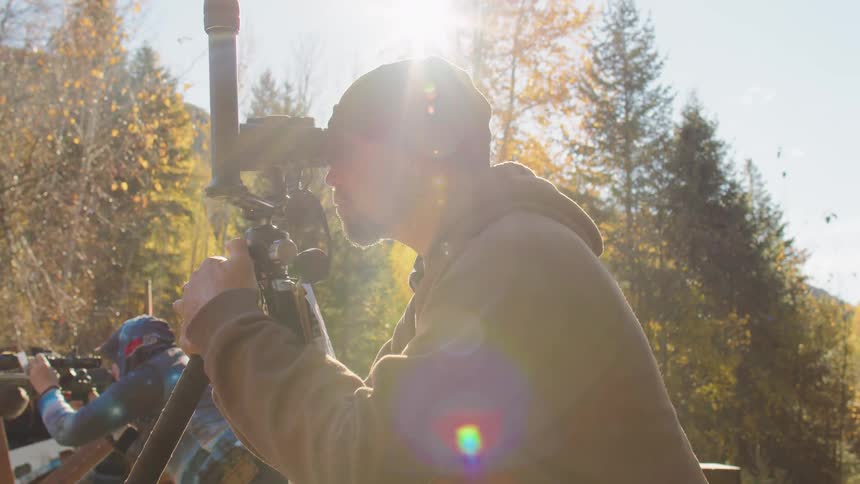 A bearded videographer in a beanie adjusts his tripod-mounted camera in the bright sunlight, creating a lens flare