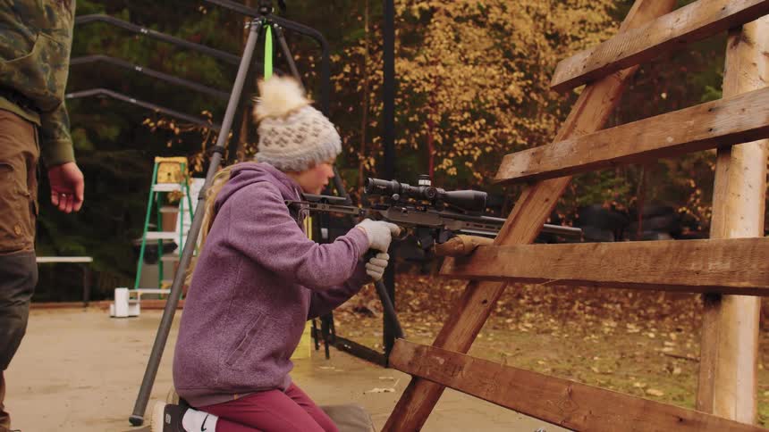 An instructor guides a young girl as she kneels and aims a high-powered, scoped rifle at an outdoor shooting range