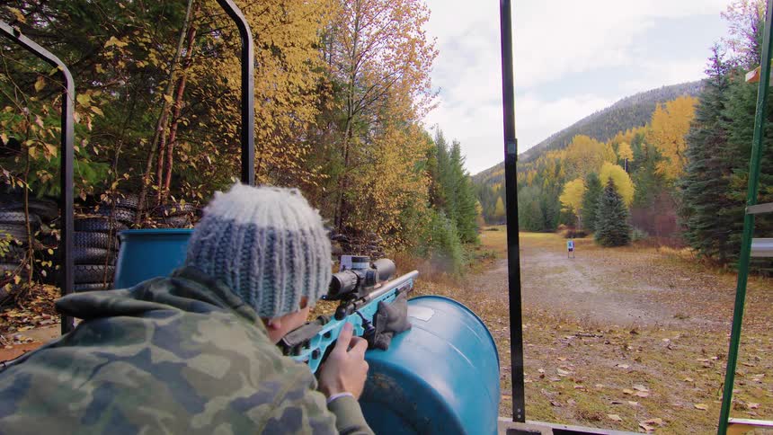 A marksman in a wool hat and camouflage jacket aims a long-range rifle with a scope at a distant target in an outdoor range during autumn