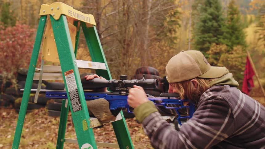 A rifleman carefully aims and fires his bolt-action rifle through an obstacle during a shooting competition in an autumn forest