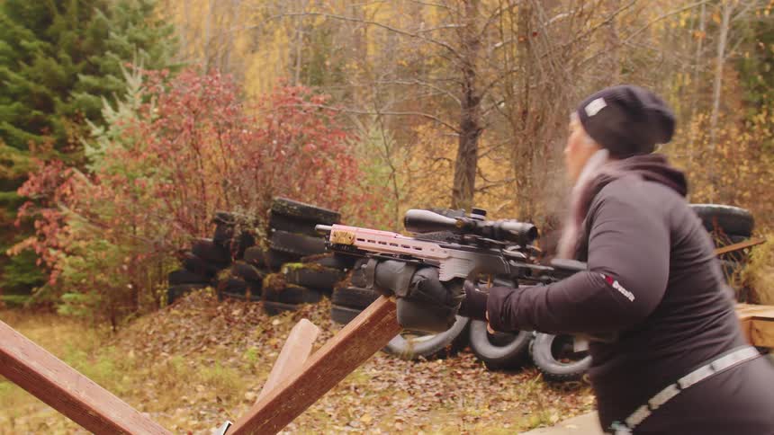 A woman in a beanie fires a high-powered rifle in slow motion at an outdoor range during a cool autumn day