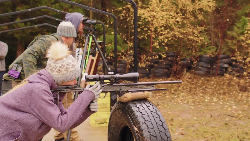 An instructor helps a young woman position and aim her long-range rifle for target practice at an outdoor range in the autumn