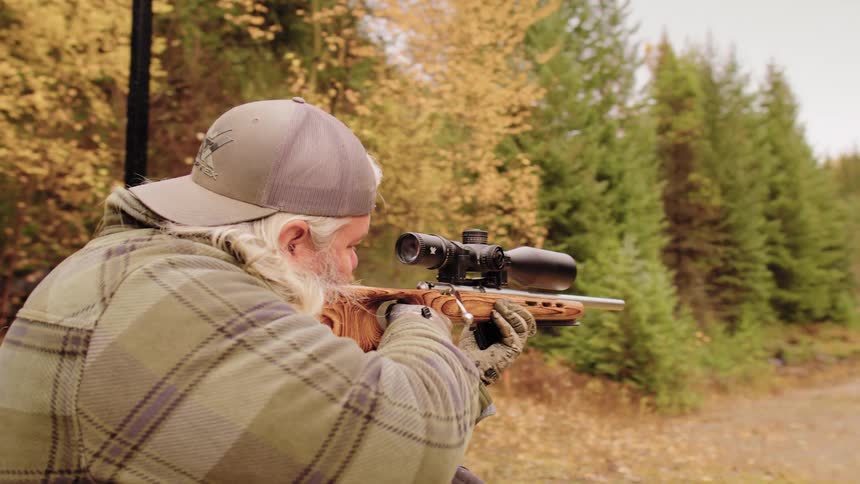 In slow motion, a man with a beard and long hair fires a scoped bolt-action rifle and reloads at an outdoor range in the fall