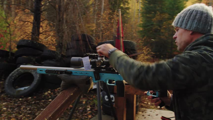 A man in a beanie aims his precision rifle and adjusts the scope at an outdoor shooting range in the forest