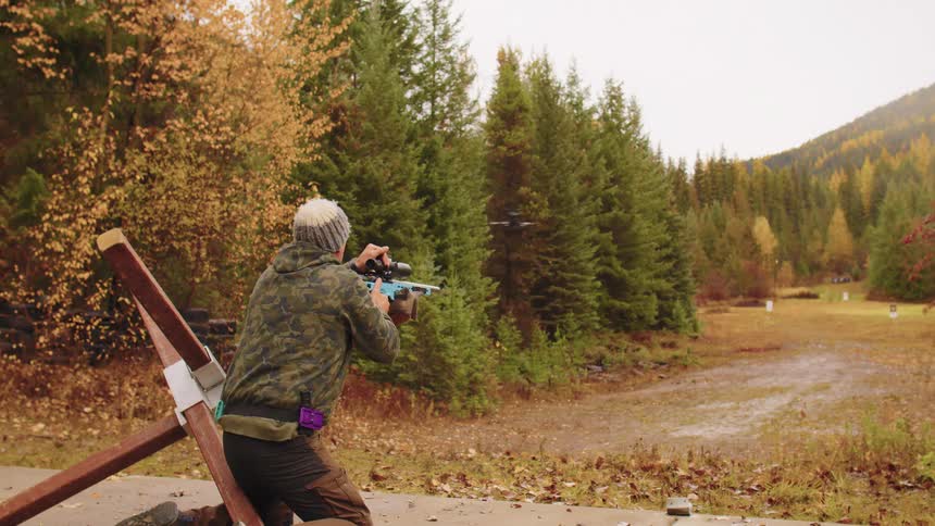 A competitive shooter in a camouflage jacket and beanie fires a rifle in slow motion from behind a wooden barricade at an outdoor range surrounded by autumn trees