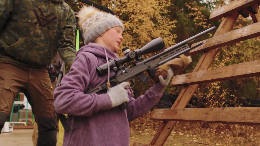 A young girl in a beanie aims and fires a bolt-action rifle from a wooden barrier during a timed shooting competition