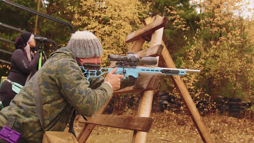 A competitive marksman aims and fires a precision rifle in slow motion from a wooden barricade at an outdoor range, with a spotter in the background