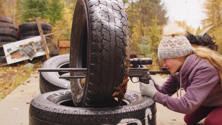 A teenage girl in a winter hat kneels and aims a scoped pistol, using a stack of tires for support during outdoor target practice in the fall