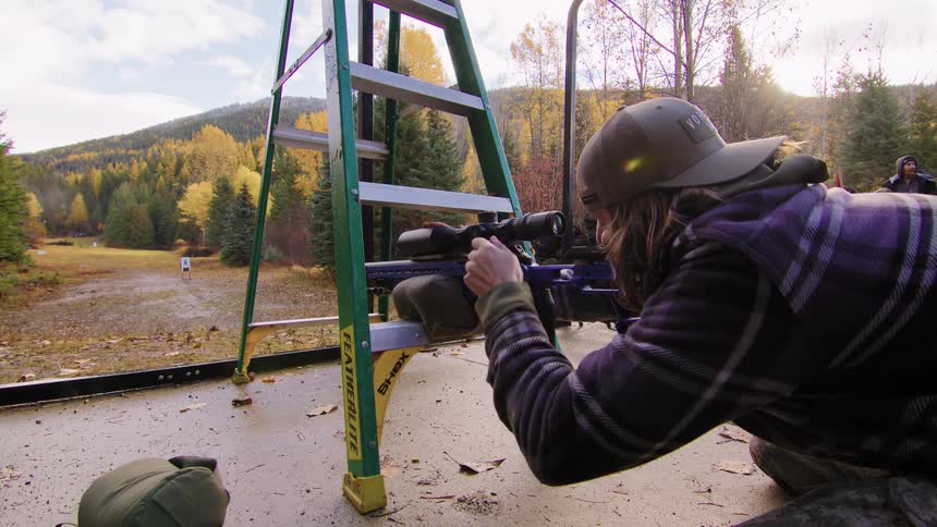 A woman fires a long-range rifle from a stabilized position on a ladder at an outdoor shooting range during autumn