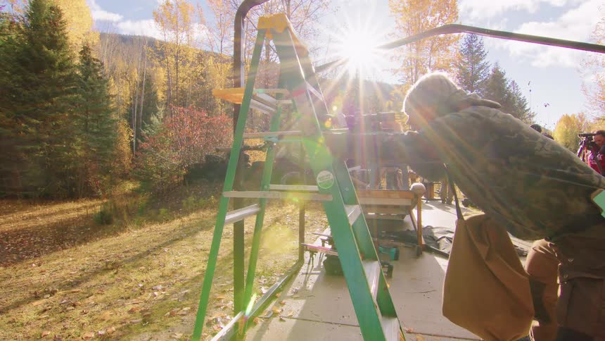 A man in camouflage takes aim and fires a sniper rifle using a ladder for support during an outdoor shooting competition on a sunny autumn day