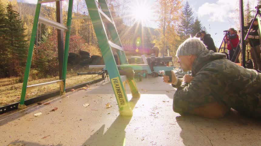 A sharpshooter in camouflage fires his rifle in slow motion from a prone position during an outdoor shooting competition