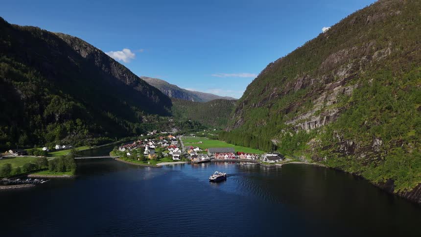 An aerial view pulls back to reveal a majestic Norwegian fjord, with a passenger ferry sailing towards a charming village surrounded by lush mountains