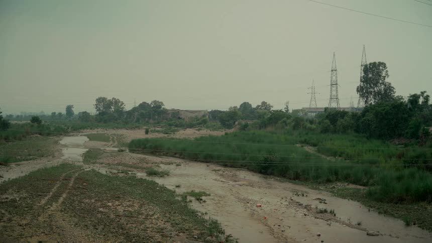 A wide, static shot of a nearly dry, muddy riverbed with lush green banks under an overcast sky, with large electricity towers in the distance
