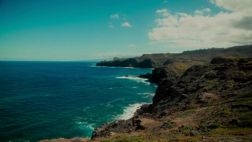A cinematic wide shot shows the turquoise ocean waves gently crashing against a rugged, rocky coastline on a beautiful sunny day