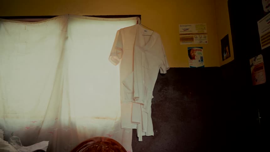 A white medical scrub uniform hangs on a hanger, backlit by a bright window in a humble, dimly lit room