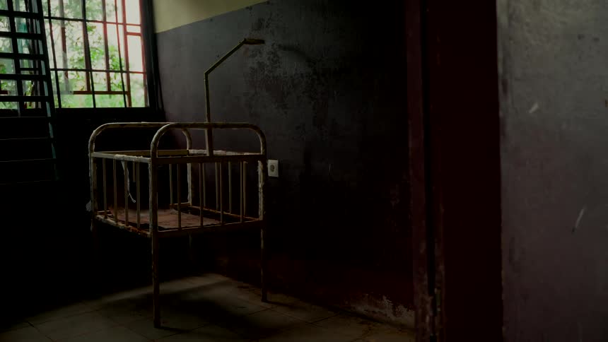 A rusty, abandoned baby crib sits alone in a dark, unsettling room of a forgotten building