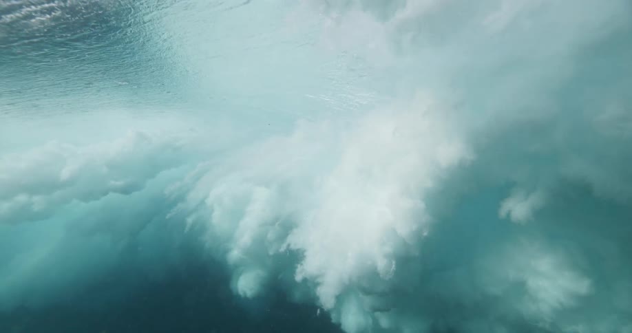 A powerful ocean wave breaks in dramatic slow motion as seen from underwater, creating a turbulent cloud of white foam