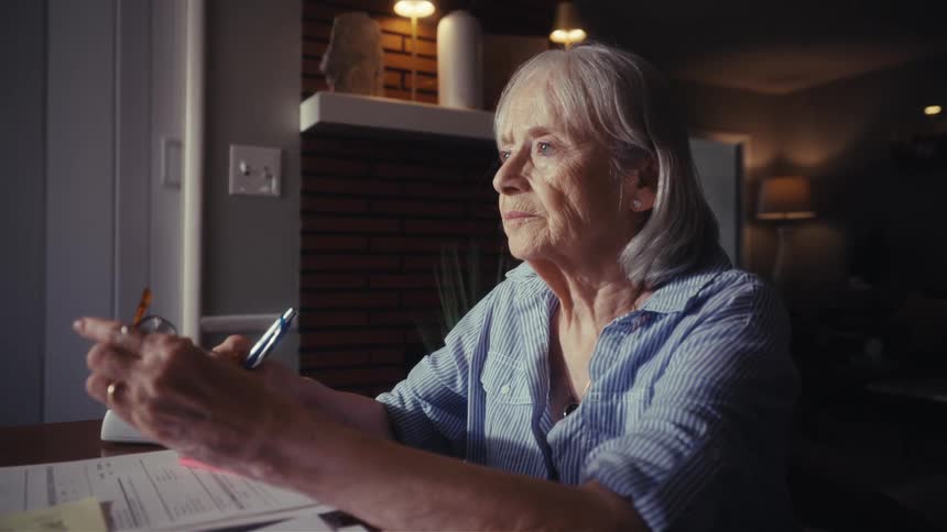 A worried elderly woman sits alone in a dimly lit room, overwhelmed by bills and financial stress as she gazes thoughtfully out the window