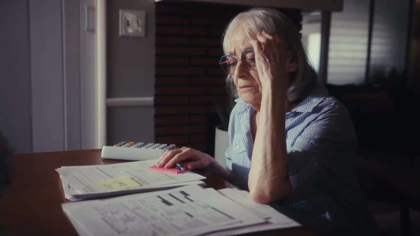 A worried senior woman with glasses sits at a table looking over bills and financial paperwork, showing signs of stress