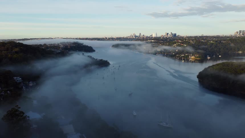 A static aerial view captures a serene, foggy morning over a wide river with boats, as the distant city skyline emerges through the mist at dawn