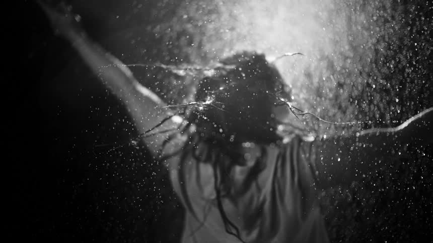 A dramatic black and white cinemagraph shows water droplets splashing over a woman with outstretched arms in super slow motion