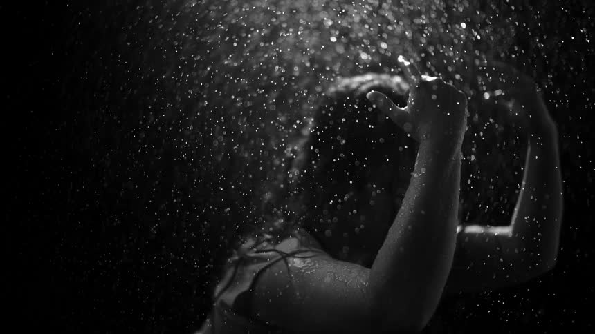 A woman with her hands in her hair is drenched by water droplets in a dramatic black and white slow-motion shot