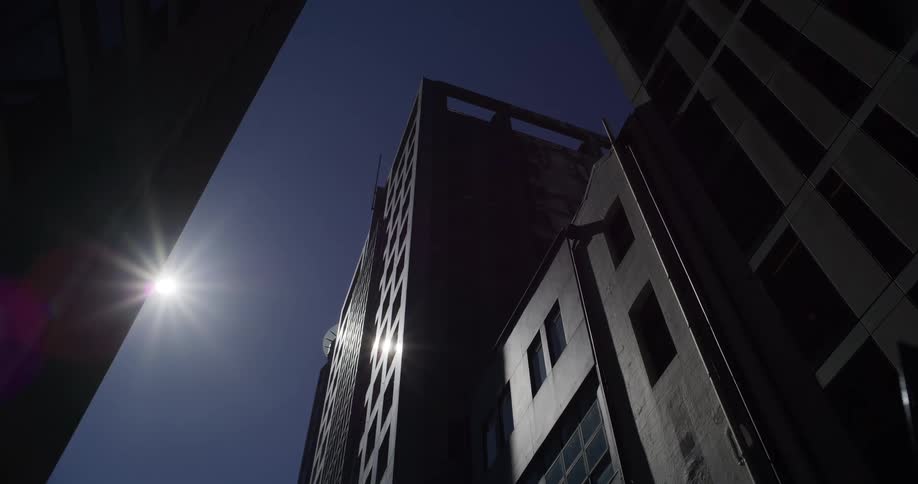 A static low-angle view shows the stark silhouettes of urban skyscrapers against a clear blue sky with a brilliant sunburst