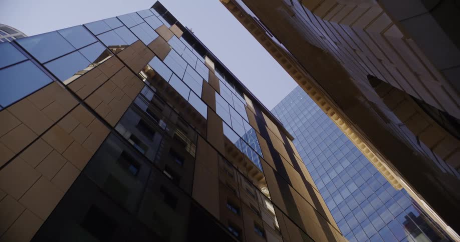 A slow zoom-out from a low-angle view of modern glass skyscrapers reflecting the clear blue sky in a city financial district