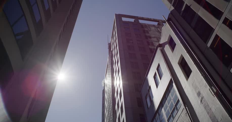 Sunlight flares over towering downtown buildings in a slow zoom-out shot looking up at the clear sky