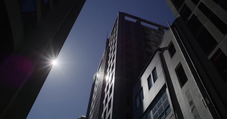 A low-angle timelapse captures the sun moving across the clear blue sky above silhouetted city skyscrapers, creating a prominent lens flare
