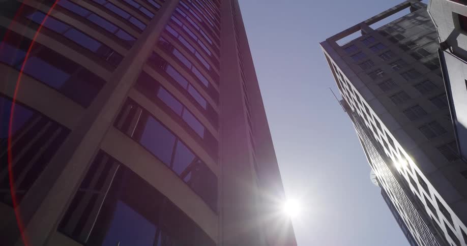 A low-angle dolly shot moves past a modern skyscraper, revealing the bright sun and a clear blue sky