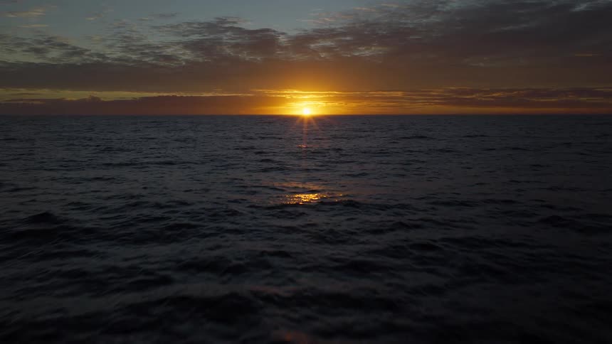 A serene aerial view flying low over the dark ocean waves towards a beautiful golden sunset on the horizon