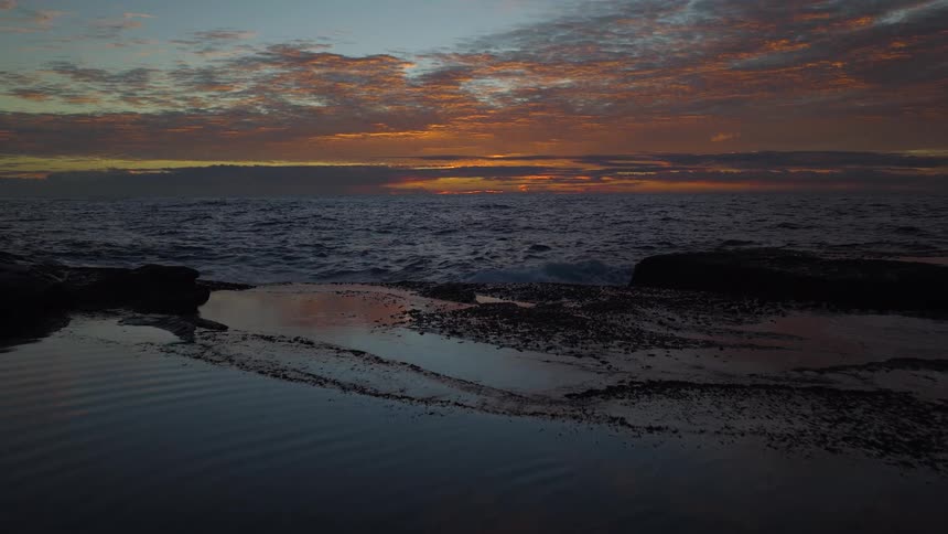 A dramatic slow motion shot of waves crashing on a rocky coast during a vibrant sunset with reflections in a tide pool