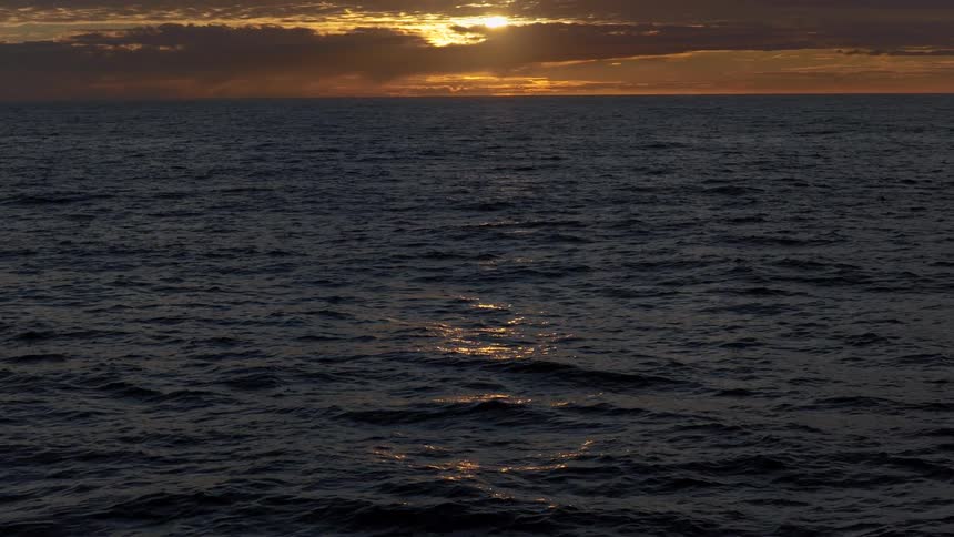 A slow tilt reveals a dramatic golden sunset reflecting on the dark, gentle waves of the open ocean