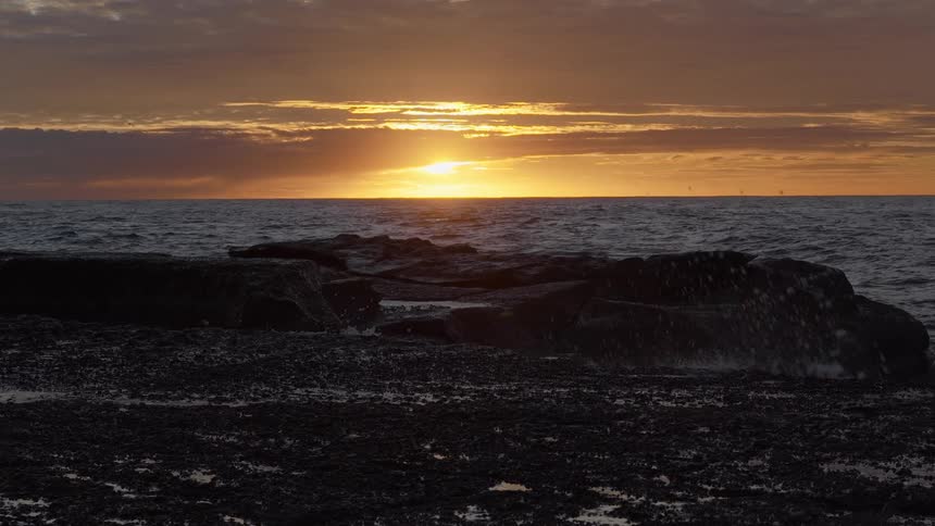 Golden sunset over the ocean highlights a dramatic slow-motion wave splash against a rocky shore