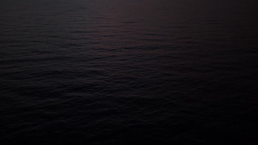 A serene aerial shot of a dark, moody ocean with gentle ripples glowing in the faint light of dusk