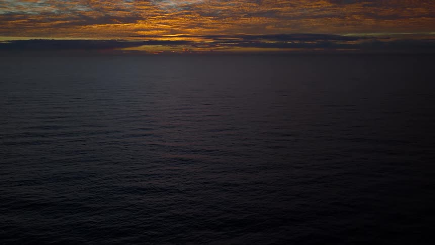 An aerial view of the dark ocean slowly tilts up to reveal a spectacular golden sunrise over the horizon