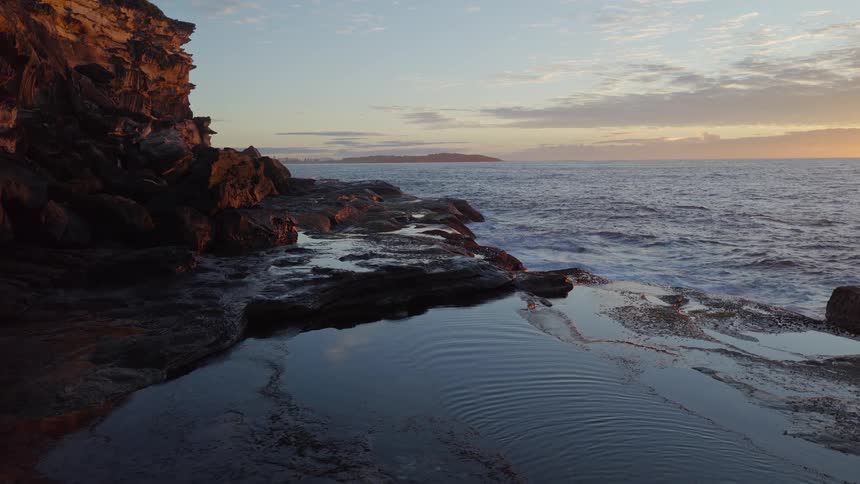 Golden sunrise light illuminates a rugged coastline as gentle ocean waves wash over the rocks and into a still pool